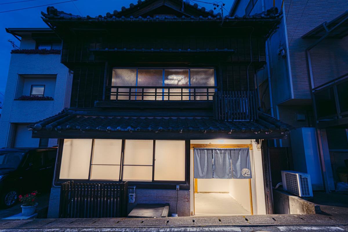 夕暮れの河崎 -湊- Minato 外観。瓦屋根の木造2階建てで、窓と暖簾のかかった玄関に明かりが灯る
