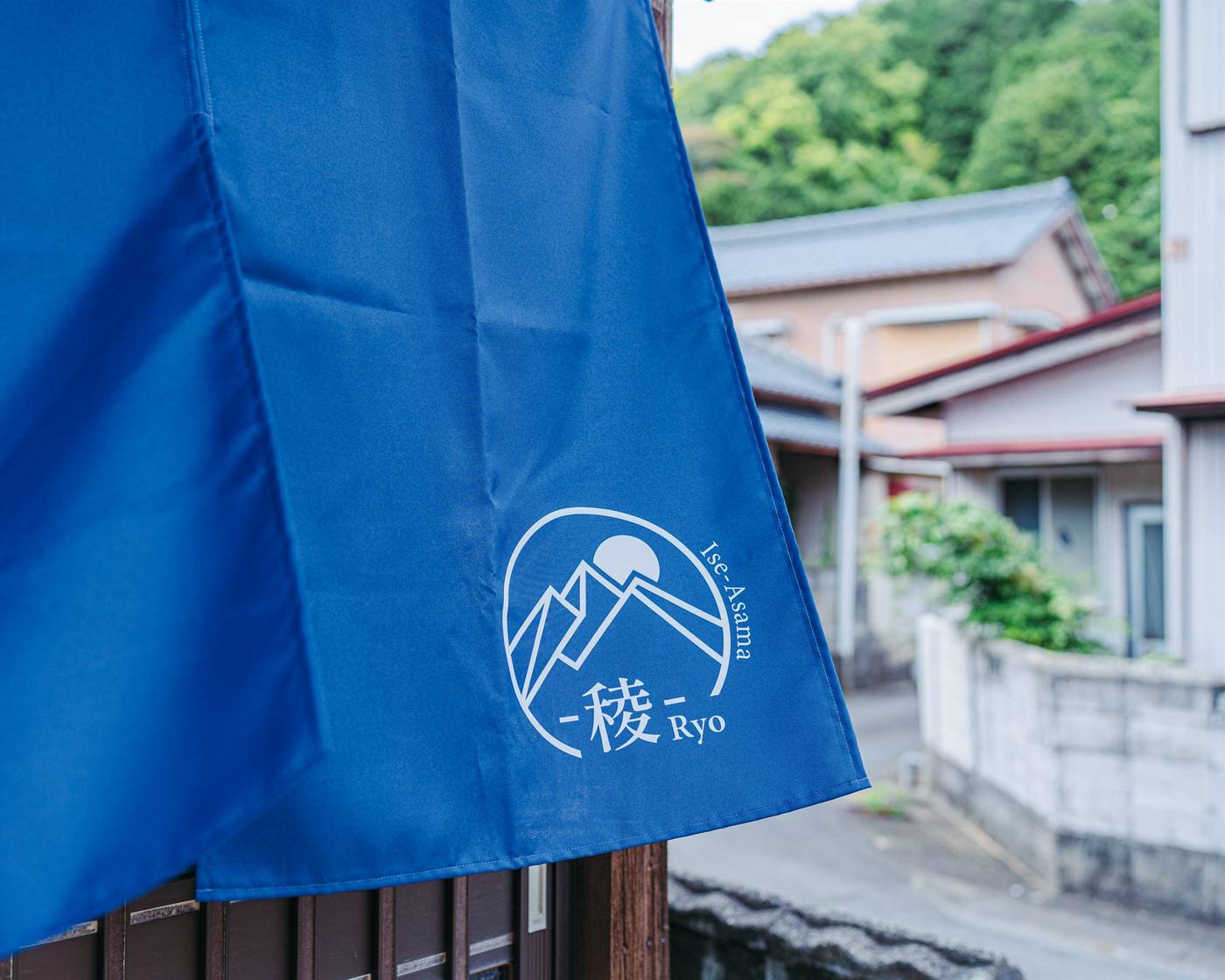 朝熊 -稜- Ryo の青い暖簾。ロゴの向こうに民家と緑の山が見える